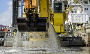 Gelbe Baggerschiff-Arme entleeren Wasser und Sedimente ins Meer. Ein riesiger Eimer schaufelt Material aus dem Wasser.