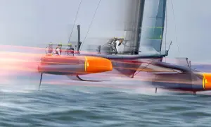 Schnelles Katamaran-Segelboot mit Besatzung, das über das Wasser gleitet. Die orangefarbenen Rümpfe und die Segel sind durch die Bewegung verschwommen, was die Geschwindigkeit des Sail GP-Rennens zeigt.