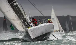 Segelboote kämpfen sich durch raue Wellen beim Fastnet Race. Ein Segler in Rot hält sich fest, während die Gischt aufspritzt.