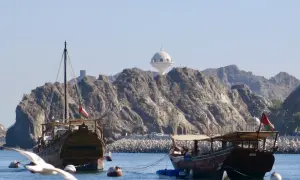 Traditionelle Dhaus-Boote im Oman Port, mit felsigen Bergen und einem weißen Kuppelgebäude im Hintergrund.