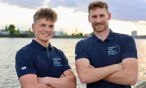 Melwin Fink und sein Teamkollege posieren lächelnd am Wasser. Sie tragen marineblaue Poloshirts mit dem Aufdruck "Around The World - Next Generation".