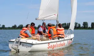 Fünf Personen in orangefarbenen Schwimmwesten genießen das Jolle segeln auf einem sonnigen See.