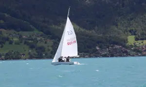 Segel reffen auf einem Boot mit drei Personen. Im Hintergrund bewaldete Hügel mit Häusern.