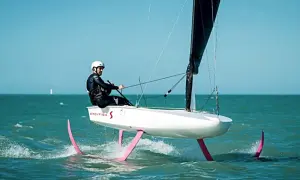 Ein Mann in Neoprenanzug und Helm steuert ein Boot mit pinken Flügeln, das über das Wasser gleitet. Das ist modernes Foil Segeln.