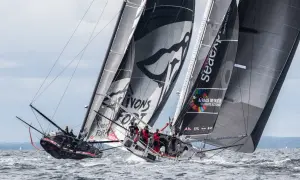 Zwei IMOCA 60 Rennyachten kämpfen auf dem Meer, Segel voller Sponsorenlogos und Botschaften für den Klimaschutz.
