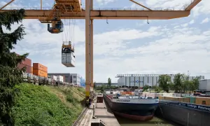 Ein Kran hebt einen Container im Hafen der Schweiz. Ein Frachtschiff liegt am Kai, umgeben von weiteren Containern und Lagerhallen.
