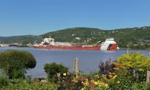 Der Frachter Paul R. Tregurtha fährt auf einem ruhigen Fluss, umgeben von grünen Hügeln und blühenden Blumen.