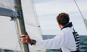 Ein junger Mann mit lockigem Haar blickt aufs Meer, während er die Hand am Mast hält. Er ist bereit, die Segel zu setzen und in See zu stechen.