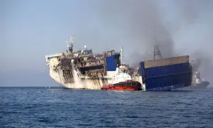Beschädigtes Schiff mit Rauch, das von einem Schlepper im Meer gezogen wird.