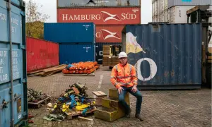Mann in Warnweste sitzt vor einem großen, blauen conex container. Stapel von roten und blauen Containern im Hintergrund.
