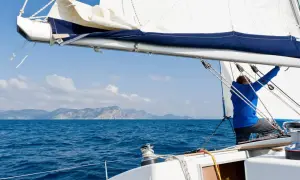 Mann in blauer Jacke trimmt das Segel eines Segelboots auf dem Meer, Berge im Hintergrund.