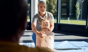 Frau zeigt an einer Übungspuppe die Herzmassage. Sie erklärt die korrekte Position für den Williamson Turn.
