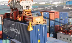 Ein Kran hebt einen blauen CMA CGM-Container. Viele weitere Container stapeln sich im Hafen, kein Container ist unberührt.