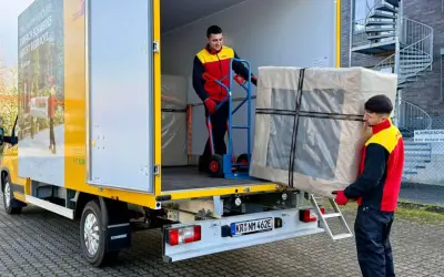 Zwei Männer laden ein großes Paket aus einem LKW. Ein Mann schiebt es mit einem Hubwagen, der andere trägt es. Maersk e-delivery macht es möglich.