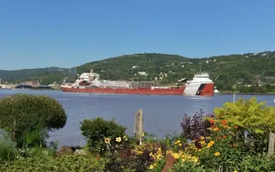 Der Frachter Paul R. Tregurtha fährt auf einem ruhigen Fluss, umgeben von grünen Hügeln und blühenden Blumen.