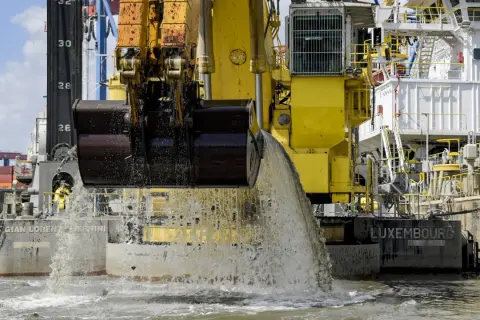 Gelbe Baggerschiff-Arme entleeren Wasser und Sedimente ins Meer. Ein riesiger Eimer schaufelt Material aus dem Wasser.