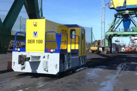 Gelbe Lokomotive "DER 100" im Riga Port, bereit für den Kohletransport unter großen Kränen.