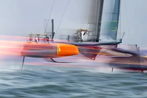 Schnelles Katamaran-Segelboot mit Besatzung, das über das Wasser gleitet. Die orangefarbenen Rümpfe und die Segel sind durch die Bewegung verschwommen, was die Geschwindigkeit des Sail GP-Rennens zeigt.