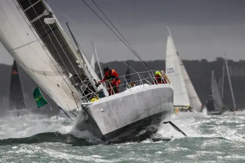 Segelboote kämpfen sich durch raue Wellen beim Fastnet Race. Ein Segler in Rot hält sich fest, während die Gischt aufspritzt.