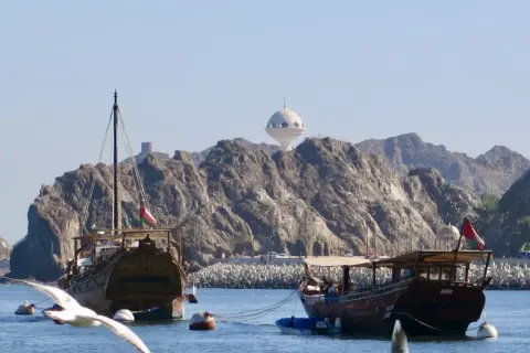 Traditionelle Dhaus-Boote im Oman Port, mit felsigen Bergen und einem weißen Kuppelgebäude im Hintergrund.