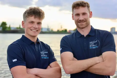 Melwin Fink und sein Teamkollege posieren lächelnd am Wasser. Sie tragen marineblaue Poloshirts mit dem Aufdruck "Around The World - Next Generation".