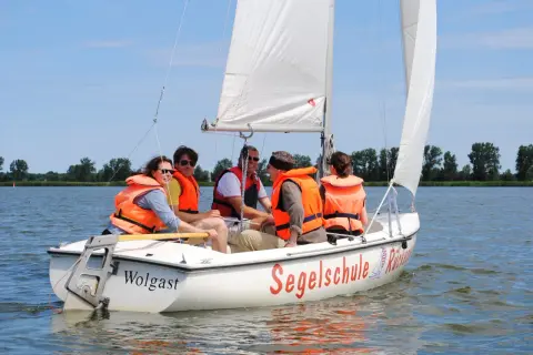 Fünf Personen in orangefarbenen Schwimmwesten genießen das Jolle segeln auf einem sonnigen See.