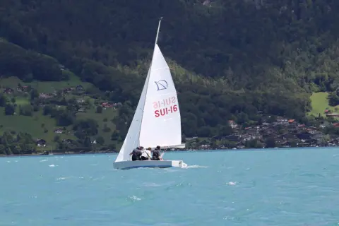 Segel reffen auf einem Boot mit drei Personen. Im Hintergrund bewaldete Hügel mit Häusern.