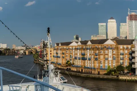 Blick auf den London Harbour mit modernen Hochhäusern und einem Flussufer mit Backsteinbauten. Ein Schiff liegt im Vordergrund.