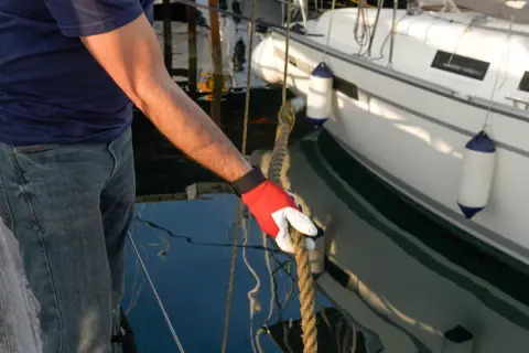 Mann mit roter Handschuh hält Seil an einem vertäuten Schiff.