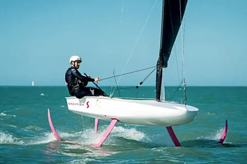 Ein Mann in Neoprenanzug und Helm steuert ein Boot mit pinken Flügeln, das über das Wasser gleitet. Das ist modernes Foil Segeln.