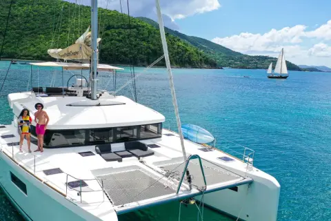 Ein Katamaran mit zwei Rümpfen, zwei Personen auf dem Deck, türkisfarbenes Wasser, grüne Hügel im Hintergrund.
