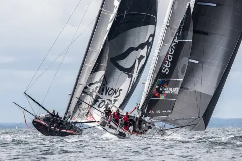 Zwei IMOCA 60 Rennyachten kämpfen auf dem Meer, Segel voller Sponsorenlogos und Botschaften für den Klimaschutz.