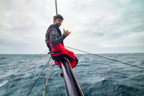 Boris Herrmann winkt lässig vom Bug seines Bootes, umgeben von rauer See und Segeltrossen.