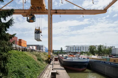 Ein Kran hebt einen Container im Hafen der Schweiz. Ein Frachtschiff liegt am Kai, umgeben von weiteren Containern und Lagerhallen.