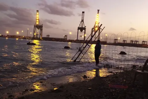Abendstimmung am Hafen von Port Sudan. Kräne leuchten im Wasser, eine Person steht am Ufer.