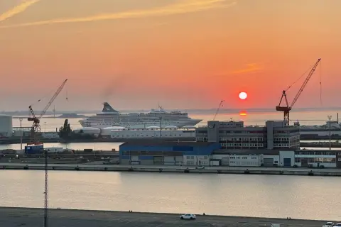 Sonnenuntergang im Hafen. Ein großes Kreuzfahrtschiff liegt vor Anker, während Kräne und Gebäude der port agency im warmen Licht erstrahlen.