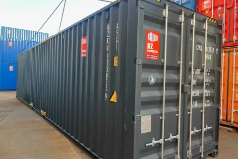 Ein grauer soc container wird von einer Kette angehoben. Daneben stehen blaue und rote Container.