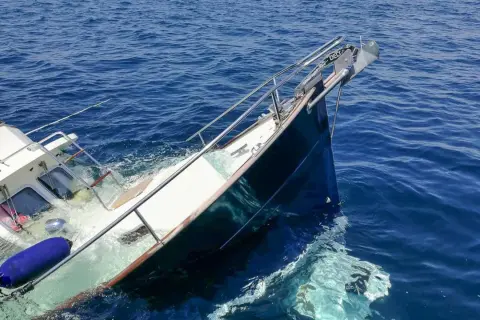 Ein sinkendes Boot neigt sich gefährlich im tiefblauen Meer. Rettungsringe und Fender treiben im Wasser.