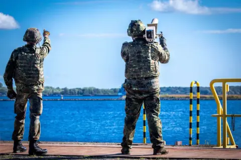 Zwei Soldaten sichern die Küste. Einer richtet eine Waffe, der andere hält ein Gerät. Maritime security in Aktion.