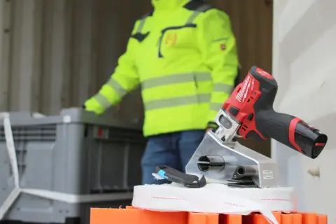 Werkzeug für Container Lashing: Ein Mann in Warnweste lädt Kisten, während ein Akkuschrauber und ein Cutter bereitliegen.