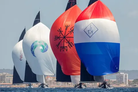 Segelboote der j class rasen über das Meer. Bunte Spinnaker mit Logos und Symbolen, im Hintergrund eine Küstenstadt.