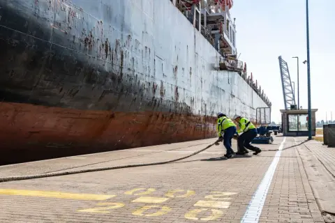Zwei Hafenarbeiter ziehen ein dickes Seil am Antwerpen Port. Ein riesiges Schiff liegt am Kai.