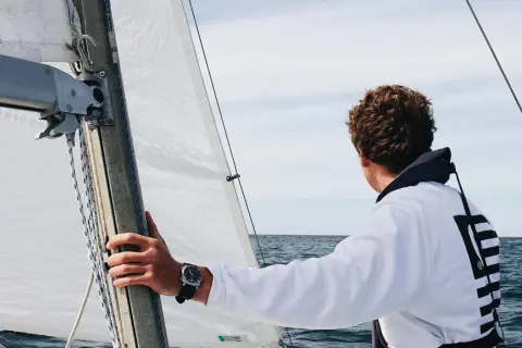 Ein junger Mann mit lockigem Haar blickt aufs Meer, während er die Hand am Mast hält. Er ist bereit, die Segel zu setzen und in See zu stechen.