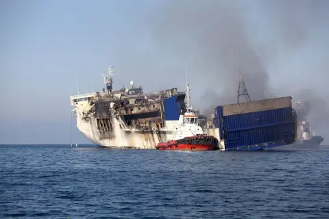 Beschädigtes Schiff mit Rauch, das von einem Schlepper im Meer gezogen wird.