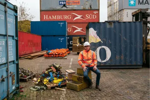 Mann in Warnweste sitzt vor einem großen, blauen conex container. Stapel von roten und blauen Containern im Hintergrund.
