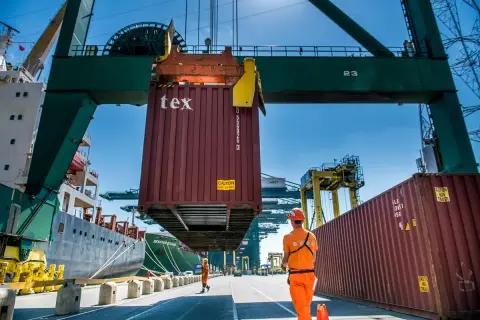 Ein Container wird im Antwerpener Hafen von einem Kran gehoben. Arbeiter in orangefarbener Kleidung überwachen die Aktion.