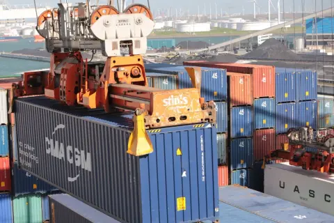Ein Kran hebt einen blauen CMA CGM-Container. Viele weitere Container stapeln sich im Hafen, kein Container ist unberührt.