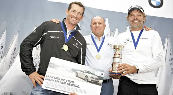 Markus Wieser mit zwei Segelkollegen und einem Pokal. Sie halten eine Broschüre für BMW Snow and Ice Training.