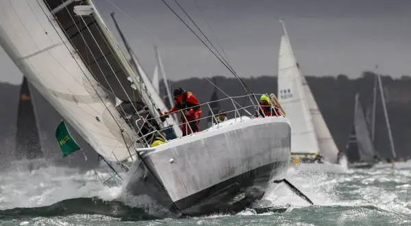 Segelboote kämpfen sich durch raue Wellen beim Fastnet Race. Ein Segler in Rot hält sich fest, während die Gischt aufspritzt.