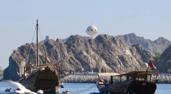Traditionelle Dhaus-Boote im Oman Port, mit felsigen Bergen und einem weißen Kuppelgebäude im Hintergrund.